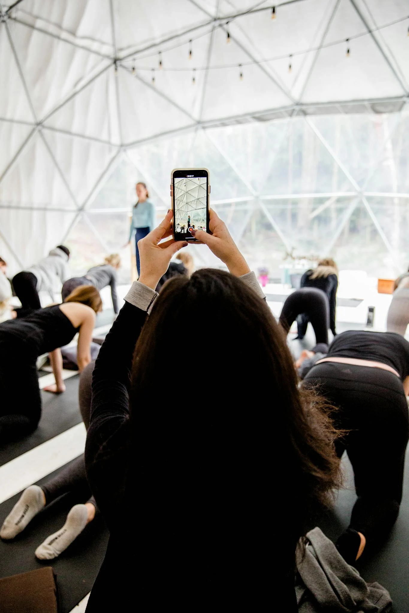 A person photographs a lively indoor yoga class using a smartphone.