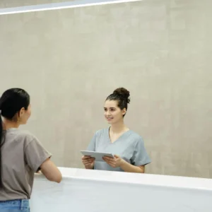 A smiling receptionist with a tablet assists a patient at a medical facility reception desk.