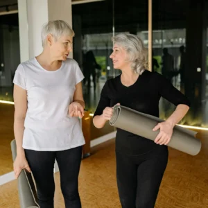 Two senior women engaging in conversation while holding yoga mats inside a fitness studio.